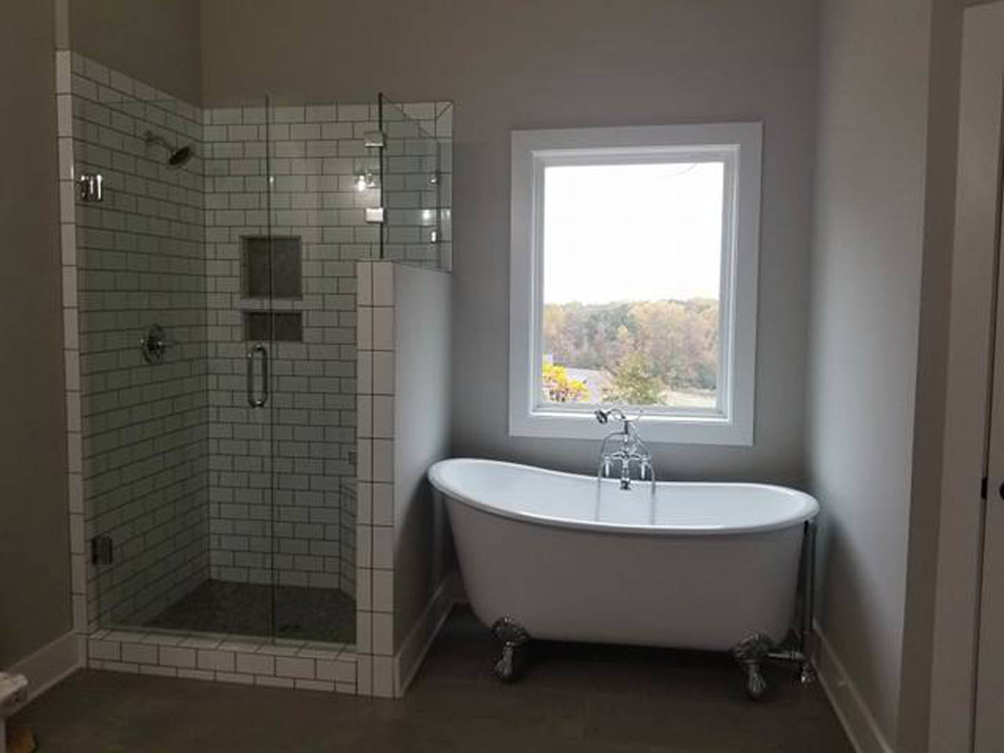 White freestanding bathtub with chrome faucet running water, glass-enclosed shower with tiled walls, large window, and light gray tile flooring