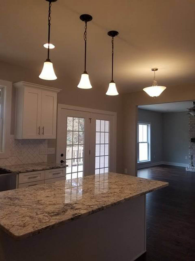 Marble countertop kitchen with white cabinetry, stainless sink, pendant lights, double glass doors, and window with white frame letting in natural light