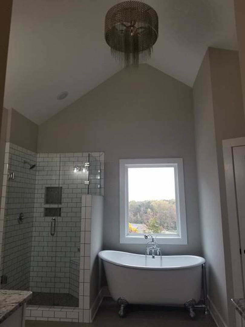 White tile bathroom featuring a freestanding bathtub with running faucet, glass-enclosed shower, large window overlooking trees, wall mirror, and ceiling fan with metal blades