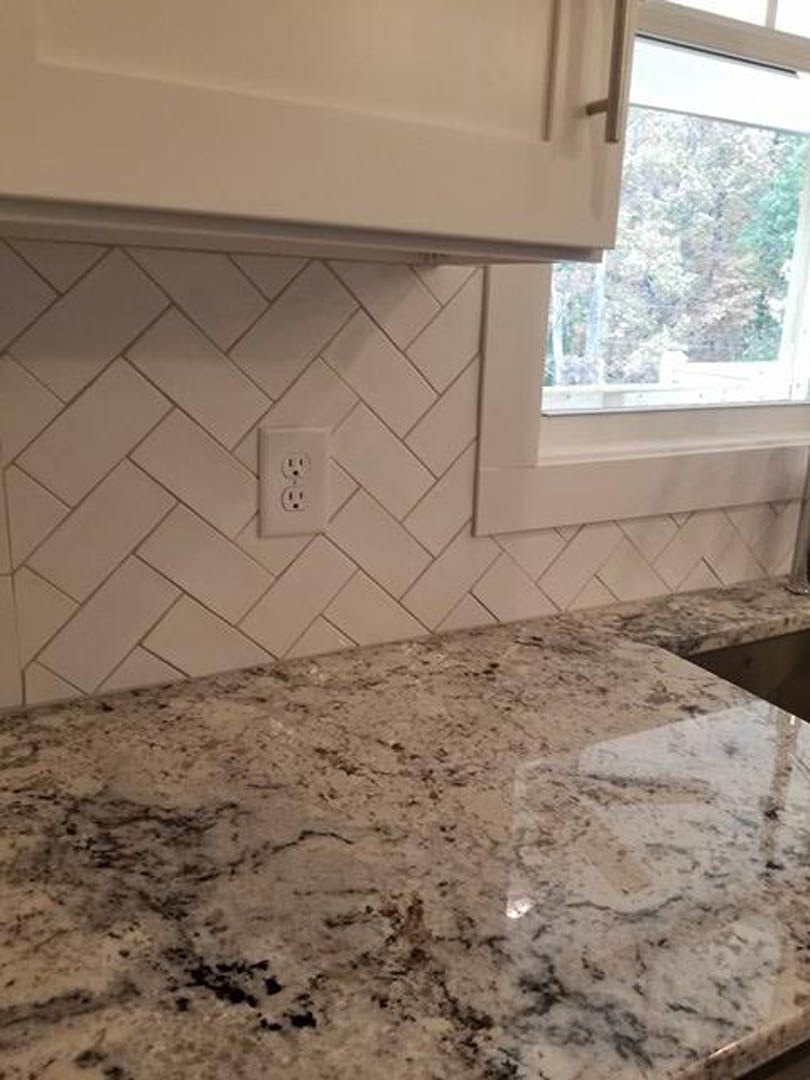 White kitchen counter with marble surface, white tile backsplash, white cabinetry with metal handles, electrical outlet, and window above sink.