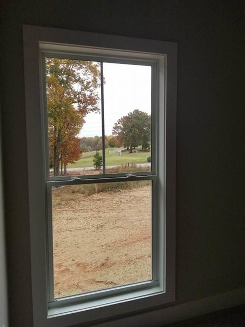 Large window framed in white trim overlooking a grassy field with scattered trees, including one with yellow leaves, and a wooden fence; indoor bench visible in foreground.