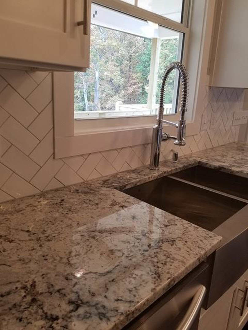 Granite kitchen countertop with stainless steel sink beneath a window showing trees outside, white cabinetry and chrome faucet visible