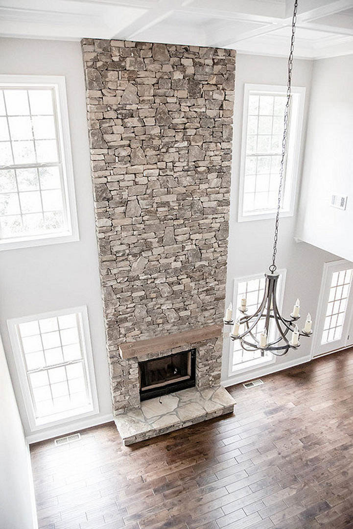 Rustic stone fireplace with candle-style chandelier hanging above, wood flooring, plaster walls, and large window in a custom home living room