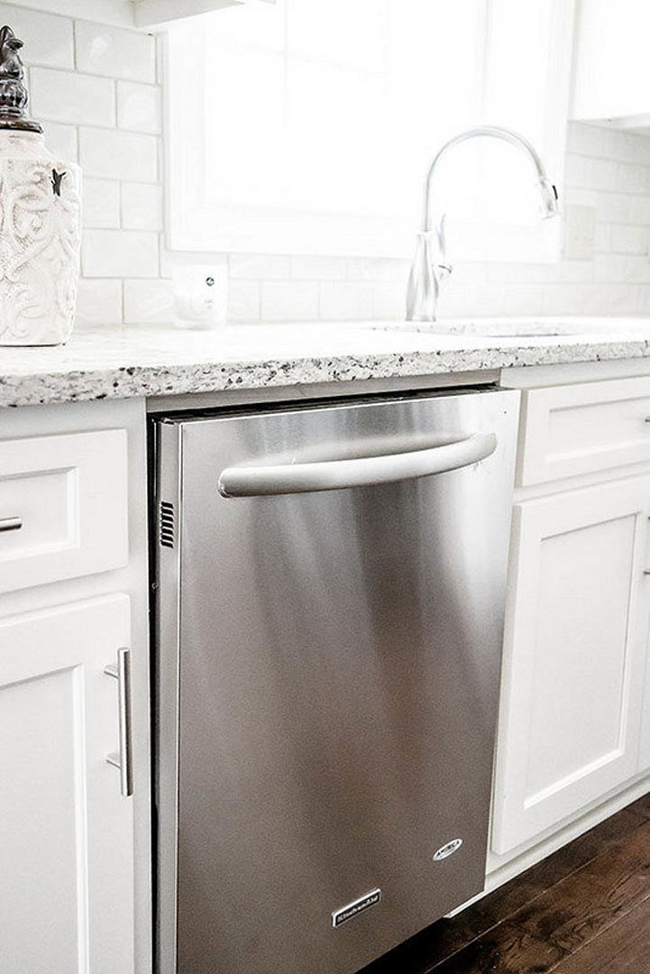 Stainless steel dishwasher beneath white quartz countertop, silver refrigerator with handle, white cabinetry, and undermount sink in modern kitchen