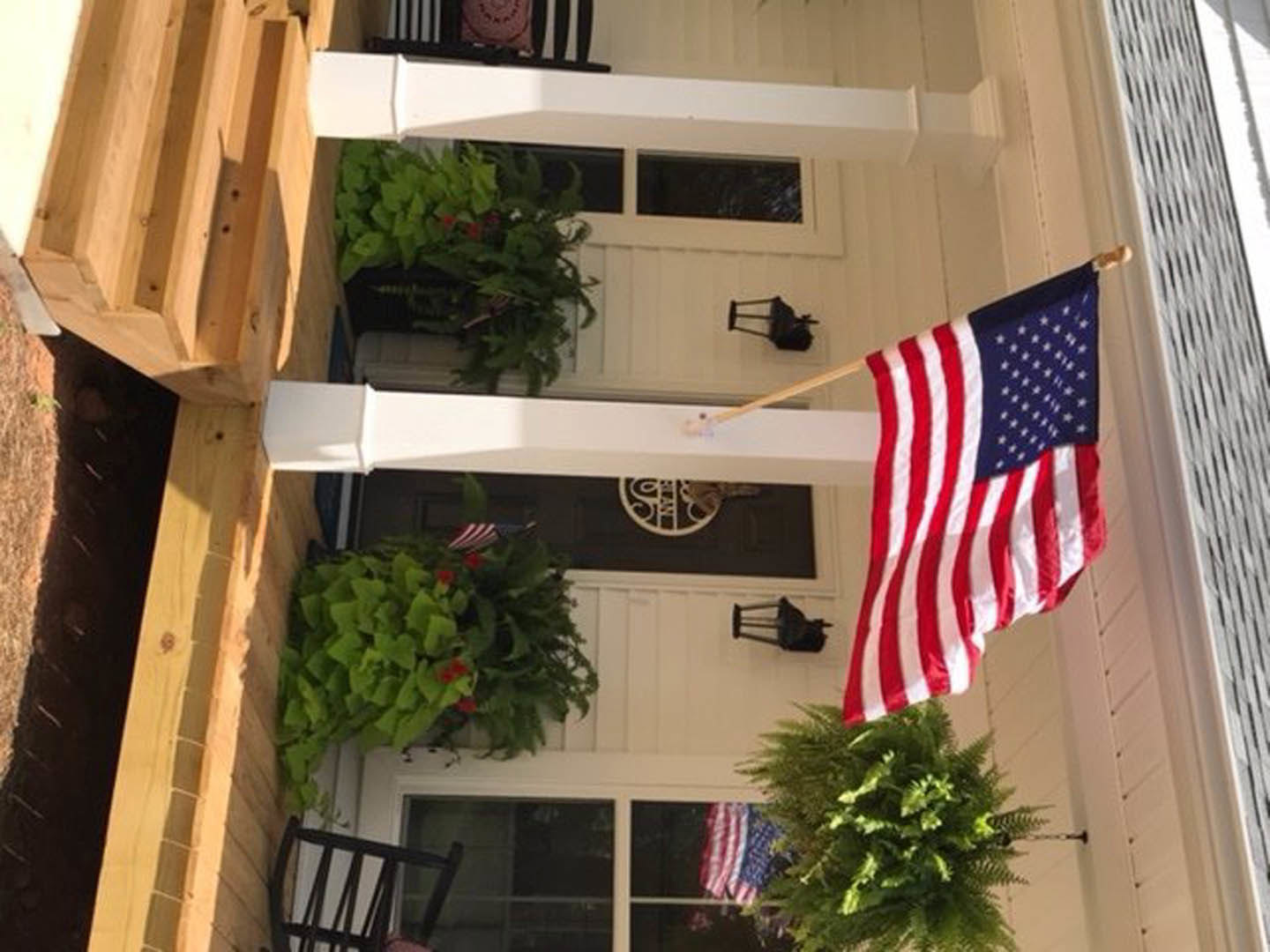 American flag hanging beside a white porch with a wooden chair, potted red flowers, and large windows
