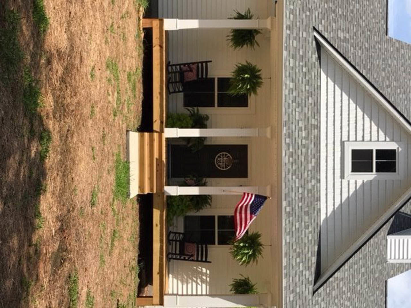 White-painted front porch with American flag hanging from railing, large windows, manicured lawn, and leafy shrubs bordering the entry.