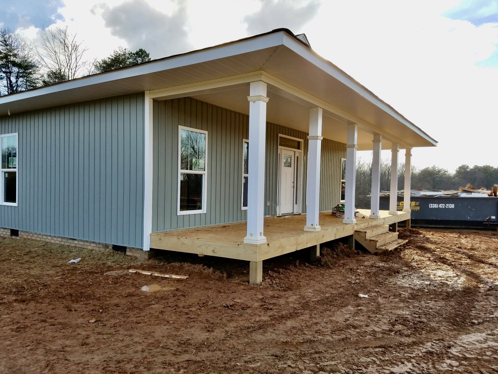 Partially built house with exposed wooden porch framing, white door with glass panel, multiple windows, and construction materials visible; trees and cloudy sky in background.