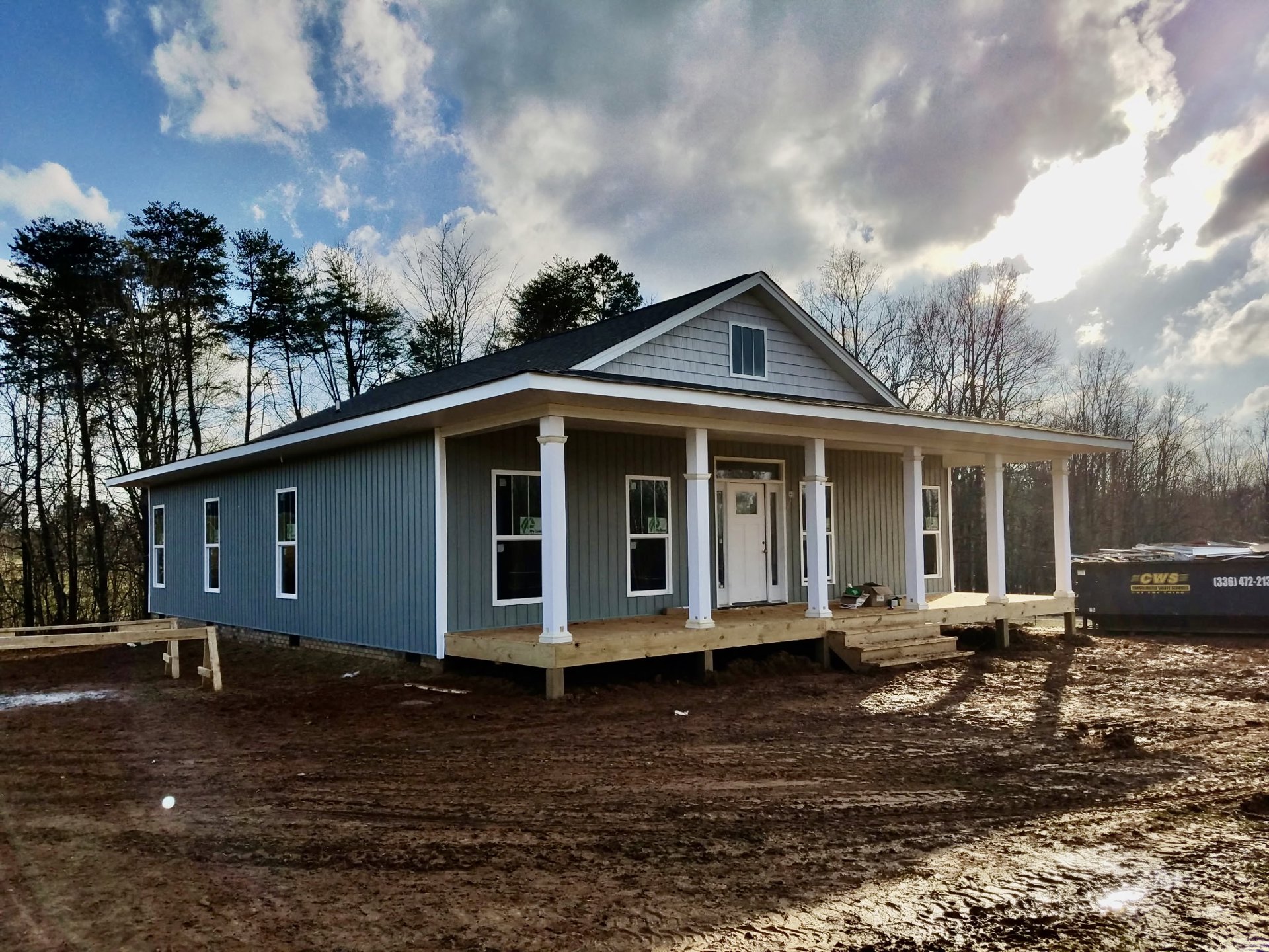 Wood-framed house under construction with covered porch, white door featuring square window, dirt yard, and trees in the background