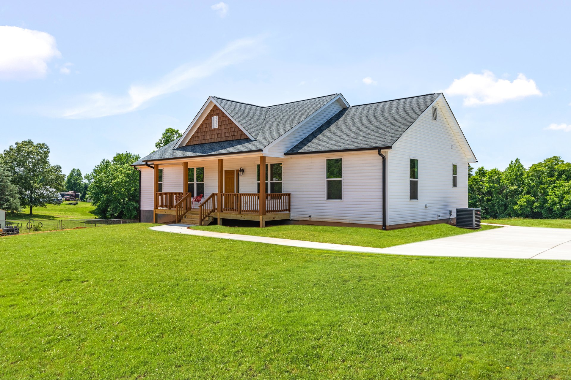 Two-story farmhouse with white siding, covered front porch with wooden railing, concrete driveway, green lawn, mature trees, and black air conditioner unit along the side.