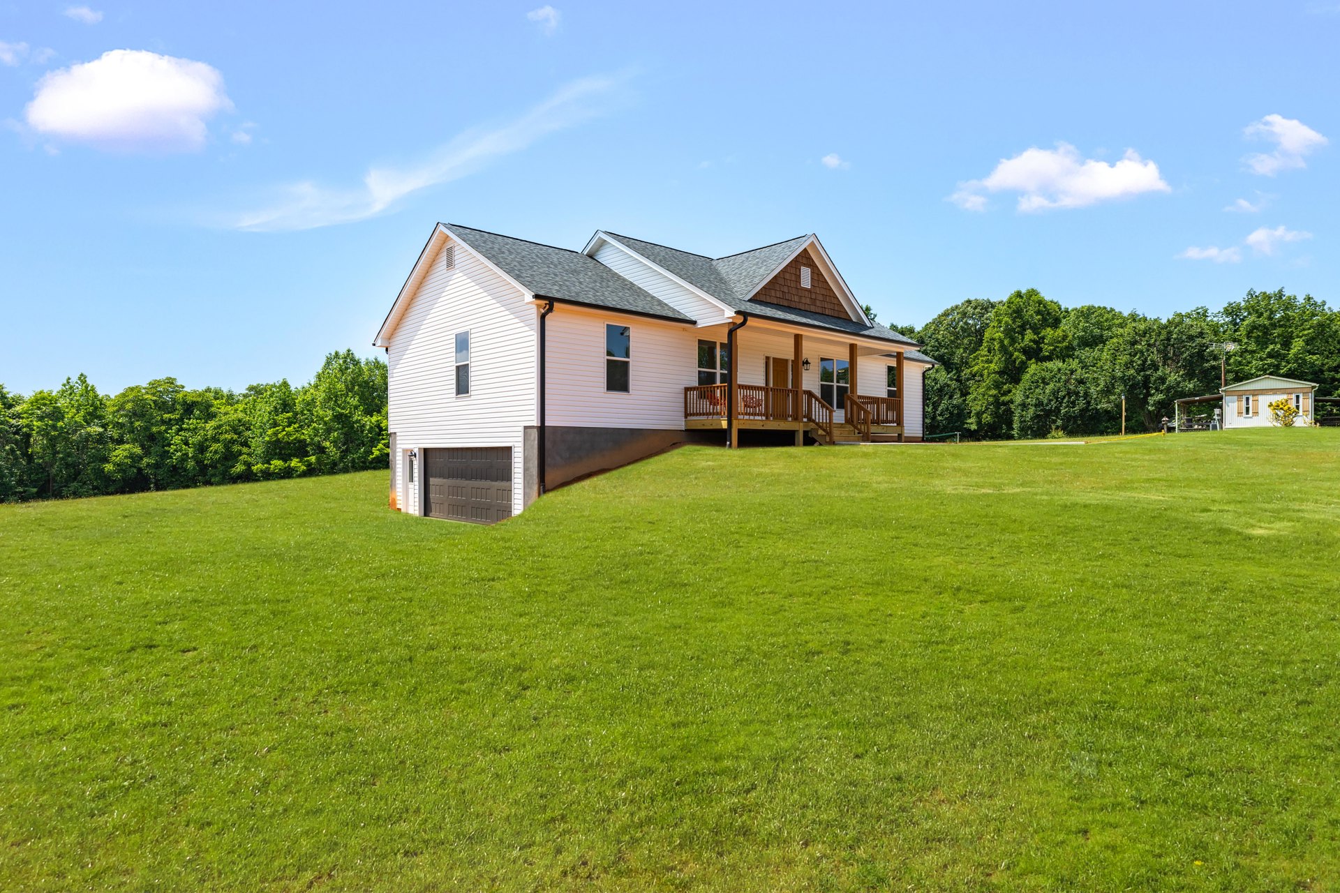 Two-story farmhouse with gray roof, attached garage, expansive green lawn, wooden deck with railing, scattered trees, and white clouds in blue sky