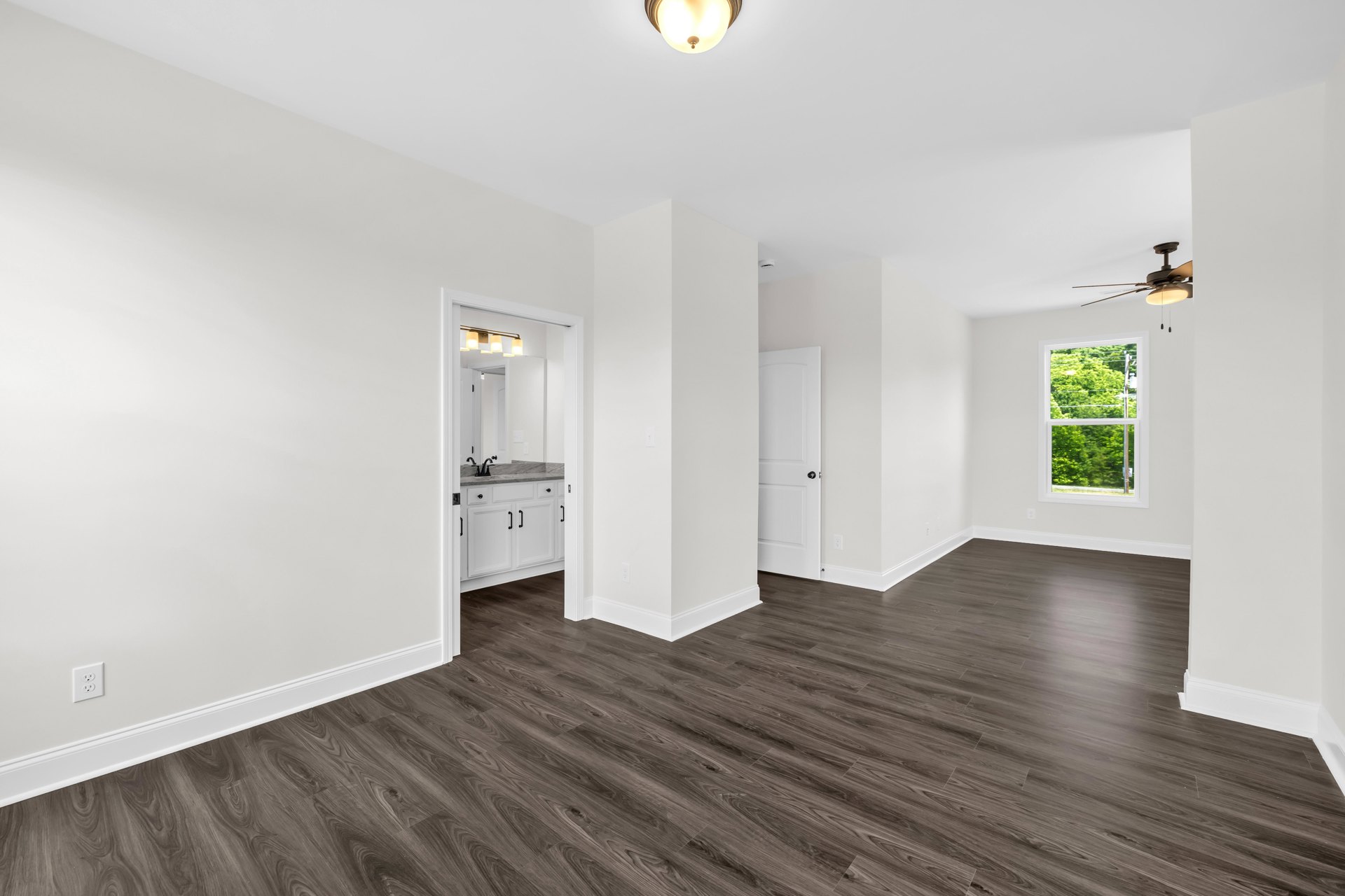 Dark wood floor in a room with white walls, white door with black knob, ceiling fan with light fixture, and window showing trees outside