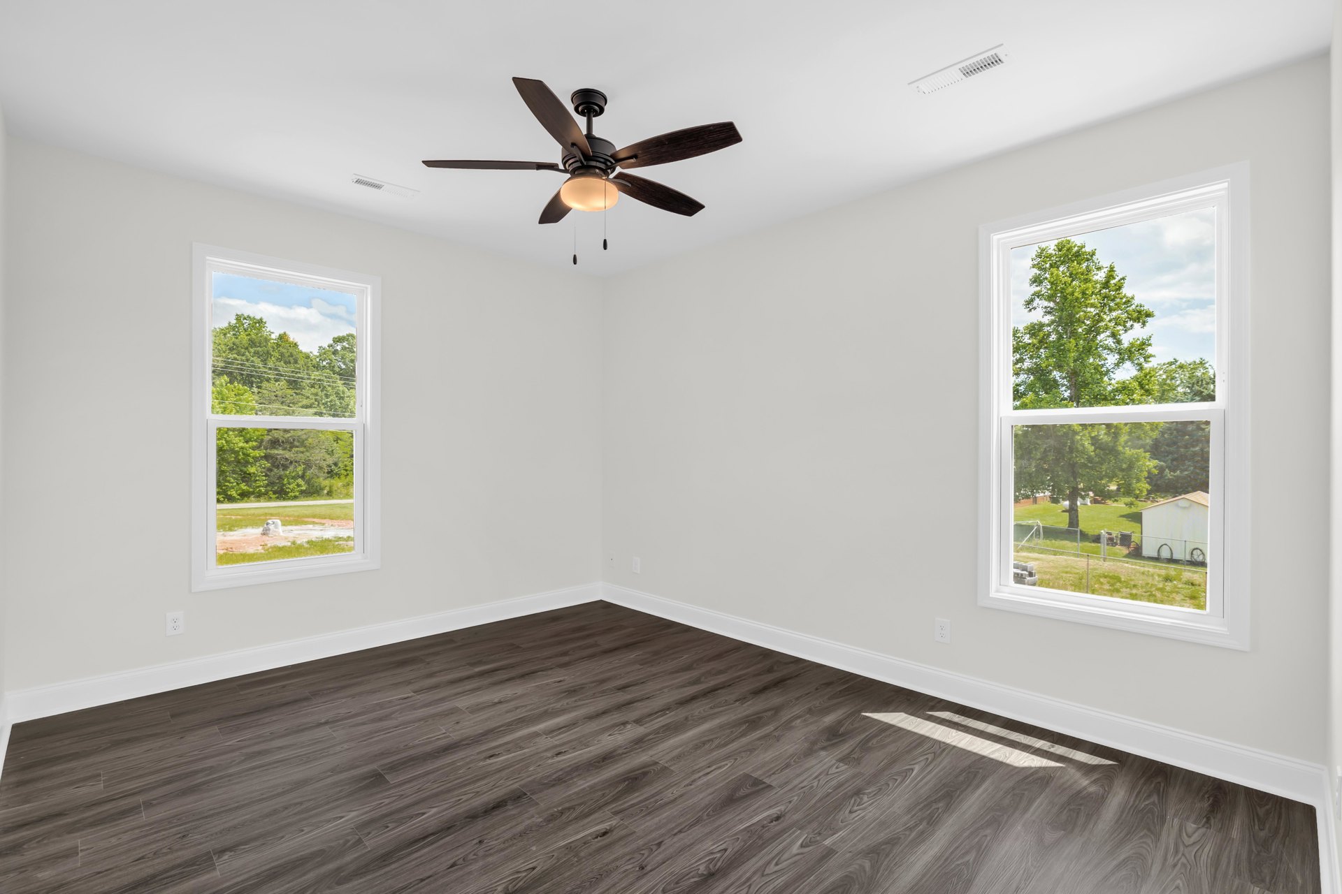 Wood flooring and white walls in a bright room with a ceiling fan, large windows overlooking green trees, and a glimpse of a white shed, fence, and bicycle outside