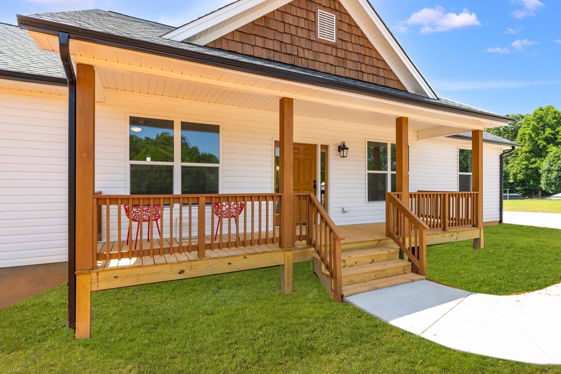 Brown-sided house with wooden porch, stairs, and concrete walkway, red chair behind fence, green lawn, white vent on exterior wall, windows visible, blue sky overhead.