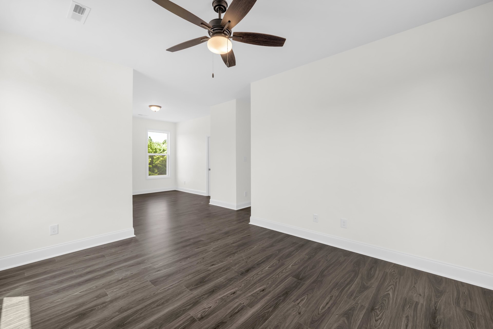 Ceiling fan with light fixture centered above hardwood floor, white plaster walls, wall vent, large window revealing leafy trees outside
