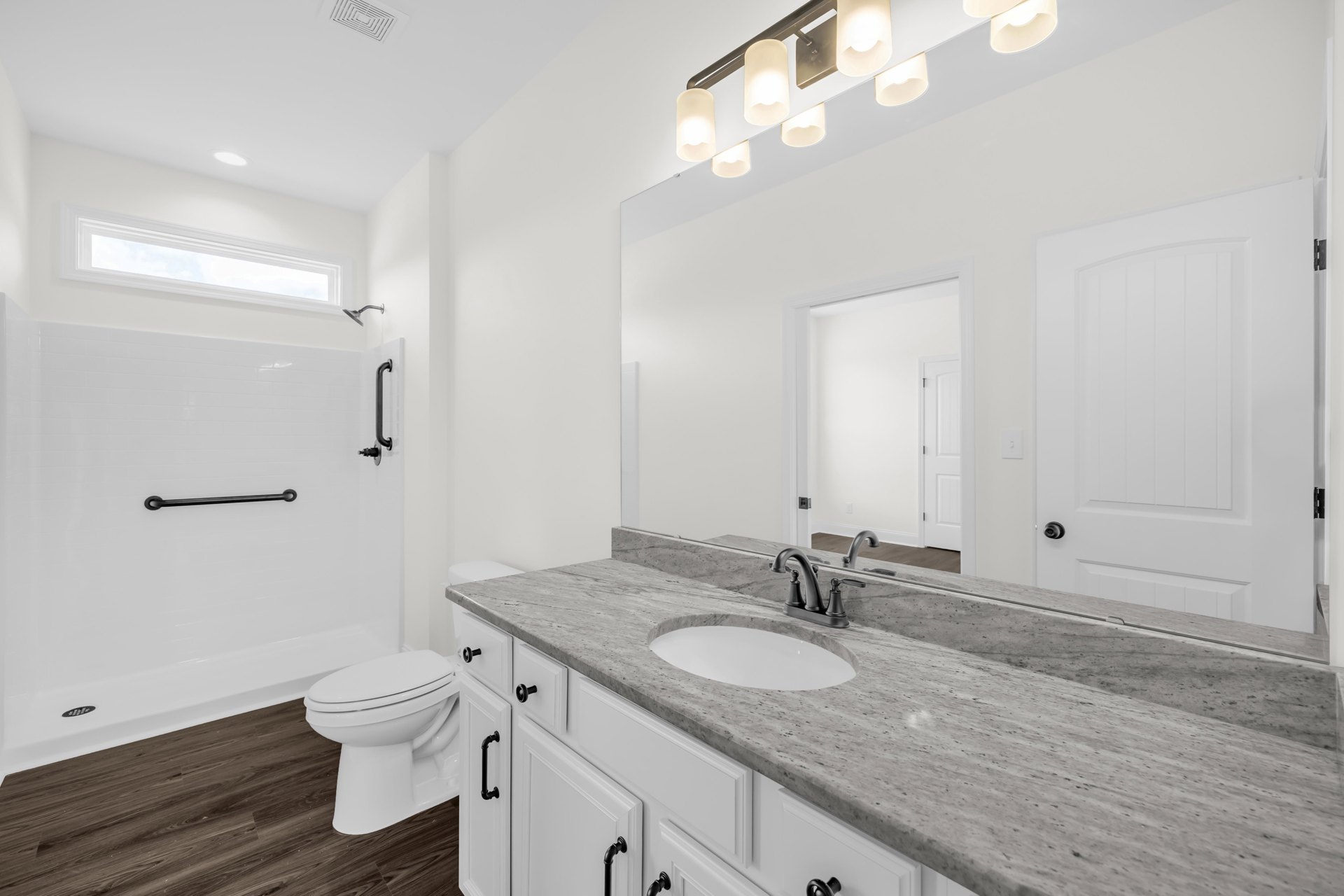 Marble countertop with undermount white sink and chrome faucet, white toilet with closed lid, wall-mounted light fixture, neutral tile walls, frameless mirror above vanity