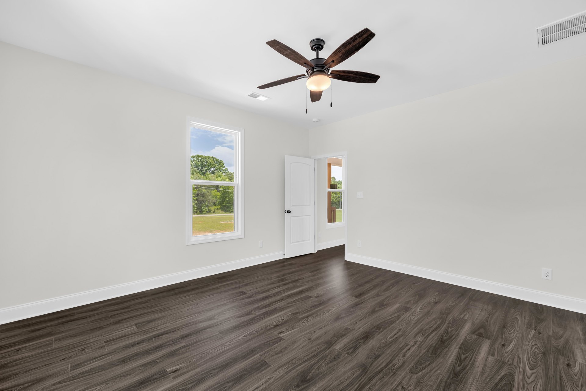 Ceiling fan with light fixture, wood flooring, white walls, window showing trees outside, white door with black knob, white ceiling vent