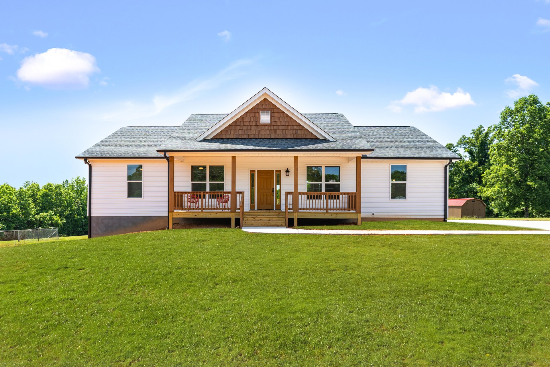 Wood-sided house with covered porch, wooden front door with glass panel, manicured lawn, leafy tree, and vented roof under partly cloudy sky