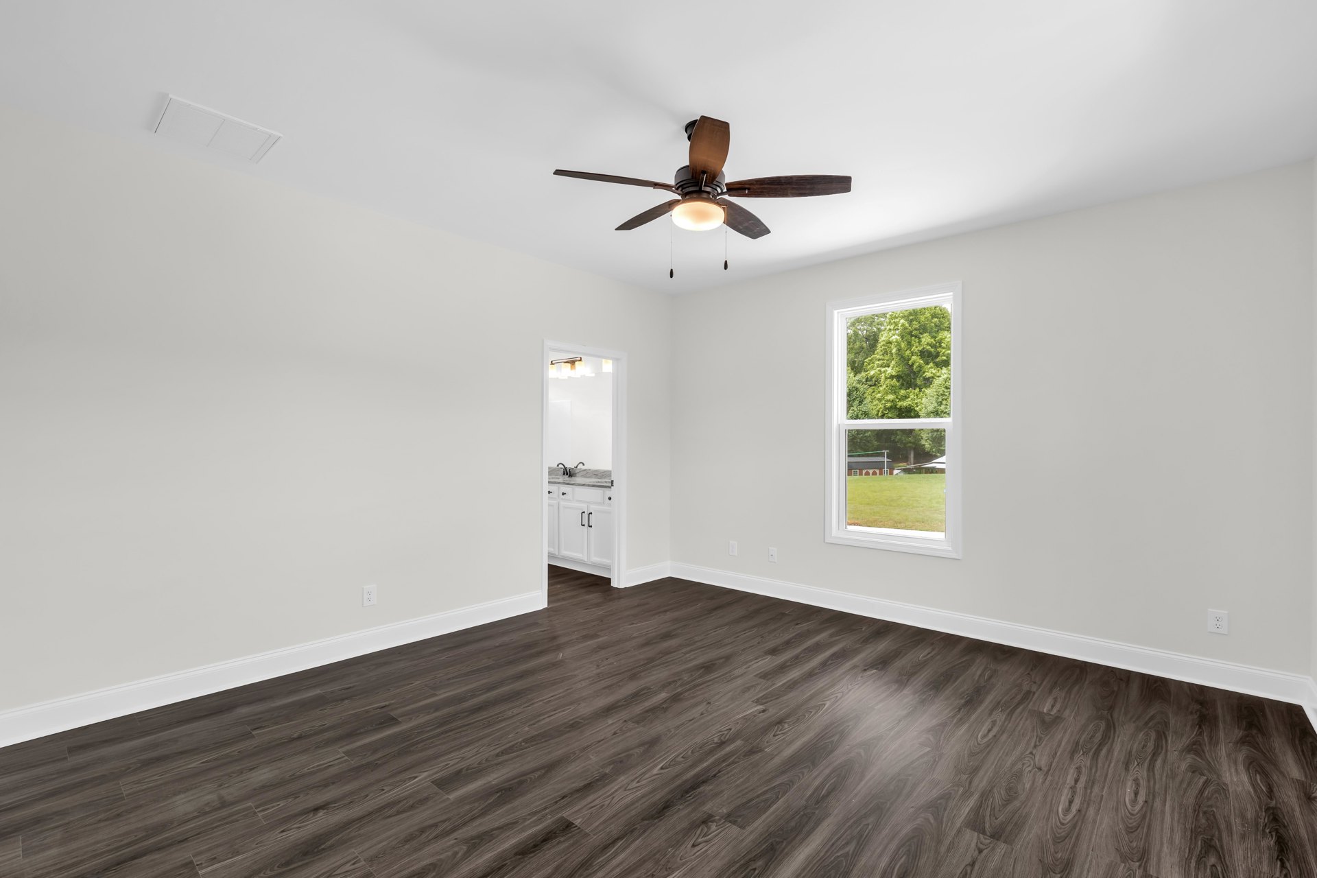Dark wood flooring in a room with white walls, ceiling fan with light, ceiling vent, marble bathroom countertop and sink, window overlooking a house and trees