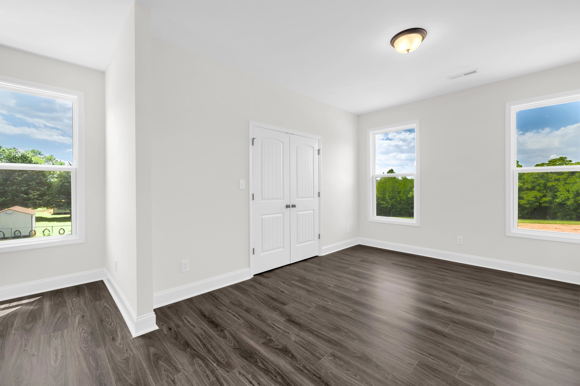 White walls and dark wood flooring in a residential room, featuring a white double door with black handles, a modern light fixture, and a window overlooking trees.