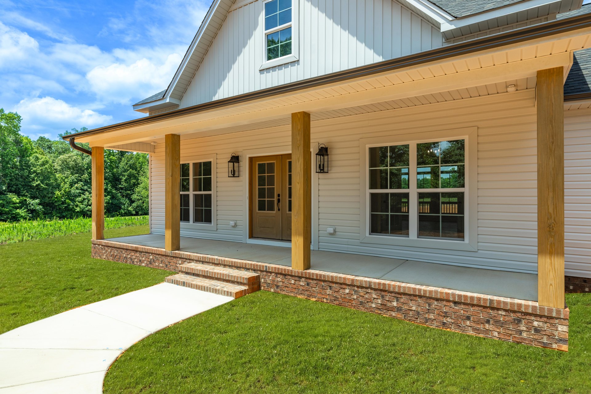 Two-story home with gray siding, covered front porch, glass-paneled door, white-framed windows reflecting trees, brick walkway and white sidewalk bordered by green lawn.