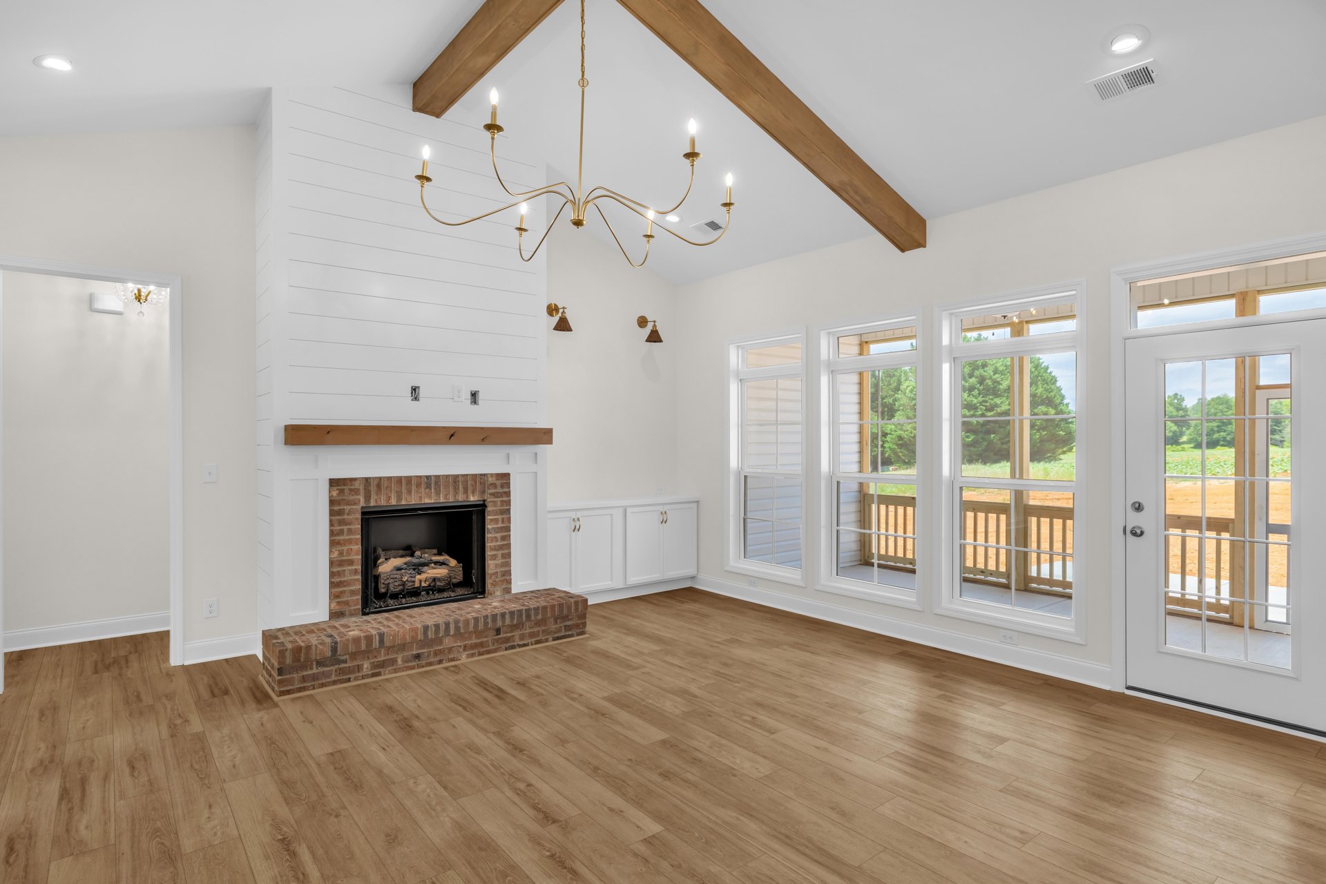 Brick fireplace with burning logs, hardwood floors, chandelier overhead, glass-paneled door opening to field view