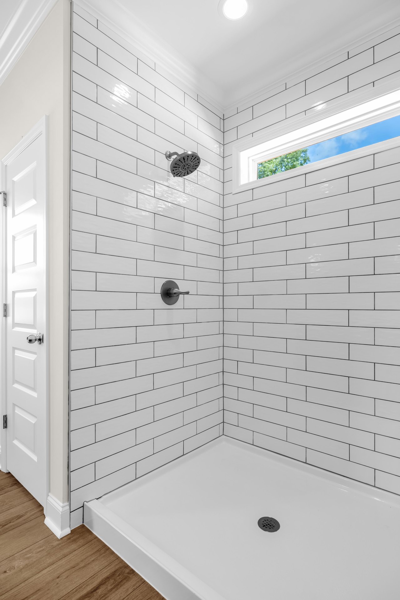 White tiled shower with chrome shower head, glass window, round drain, and close-up views of a metal knob and recessed ceiling light.