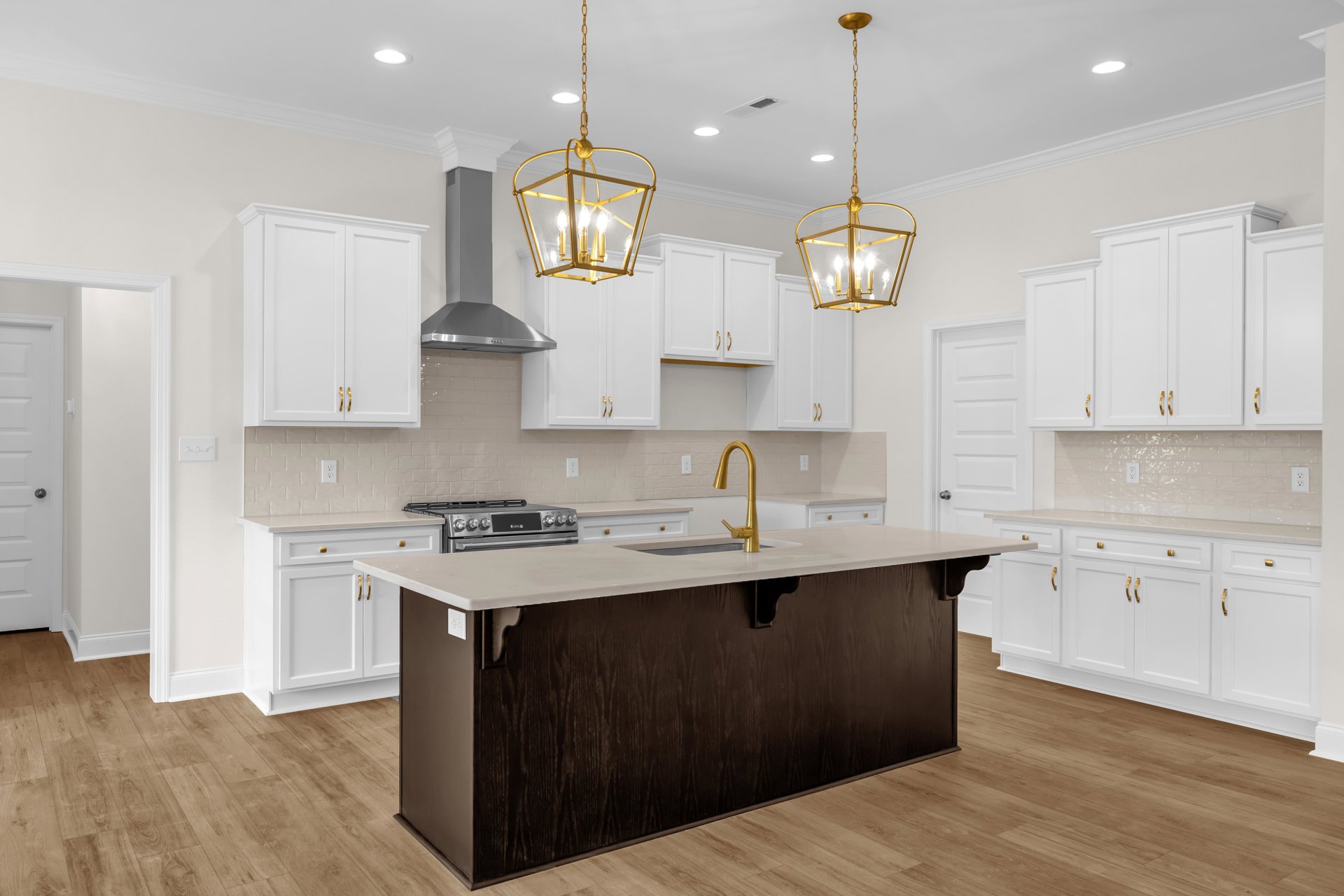 White kitchen cabinets and drawers, large central island with built-in sink, gold chandelier hanging from ceiling, light hardwood flooring, stainless steel appliances, white stone