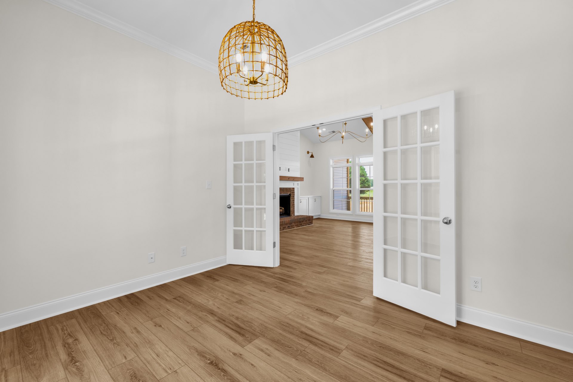 Wood flooring, white paneled doors with glass panes, gold three-light chandelier, brick fireplace wall, white plaster walls, decorative molding