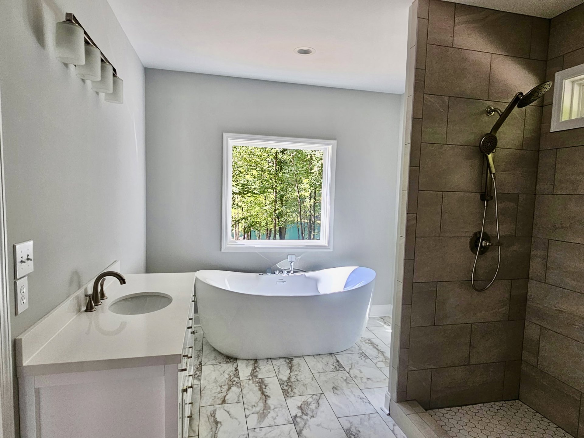 Bathroom with tiled walls, freestanding tub, single sink vanity, broken faucet, window overlooking trees, shower head with hose, white door featuring vent