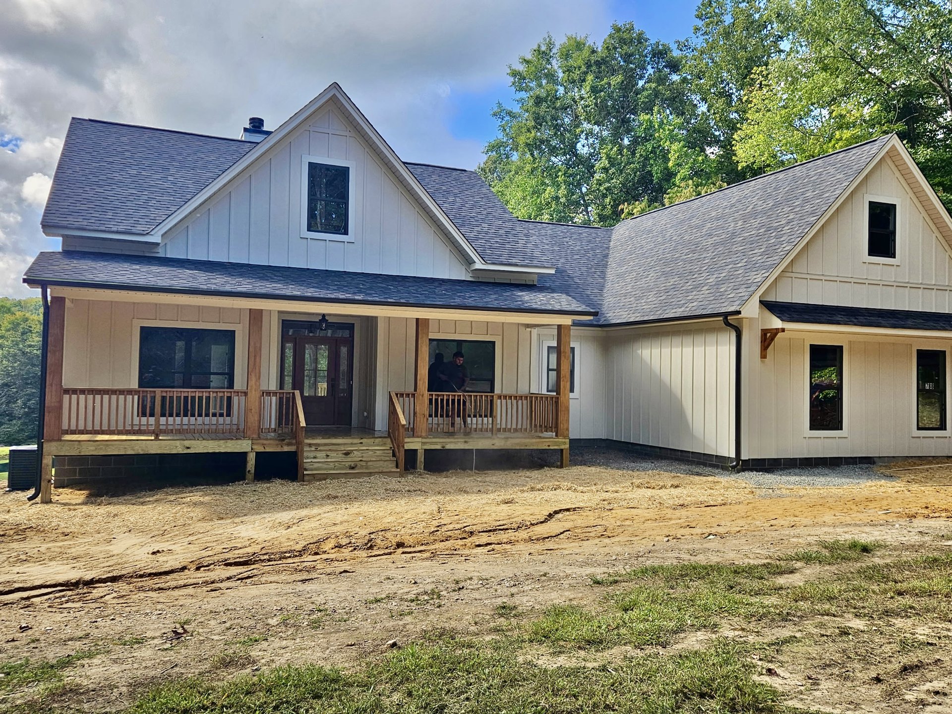 Two-story house with white siding, black-framed windows, glass-paneled front door, covered porch, dirt path leading to entrance, wooden fence in foreground, man standing on porch