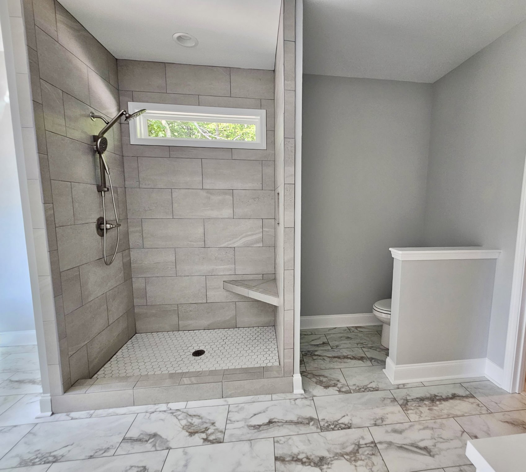 White tile bathroom with marble floor, walk-in shower featuring chrome shower head and hose, white toilet against plaster wall, window with white frame, floor drain near shower.