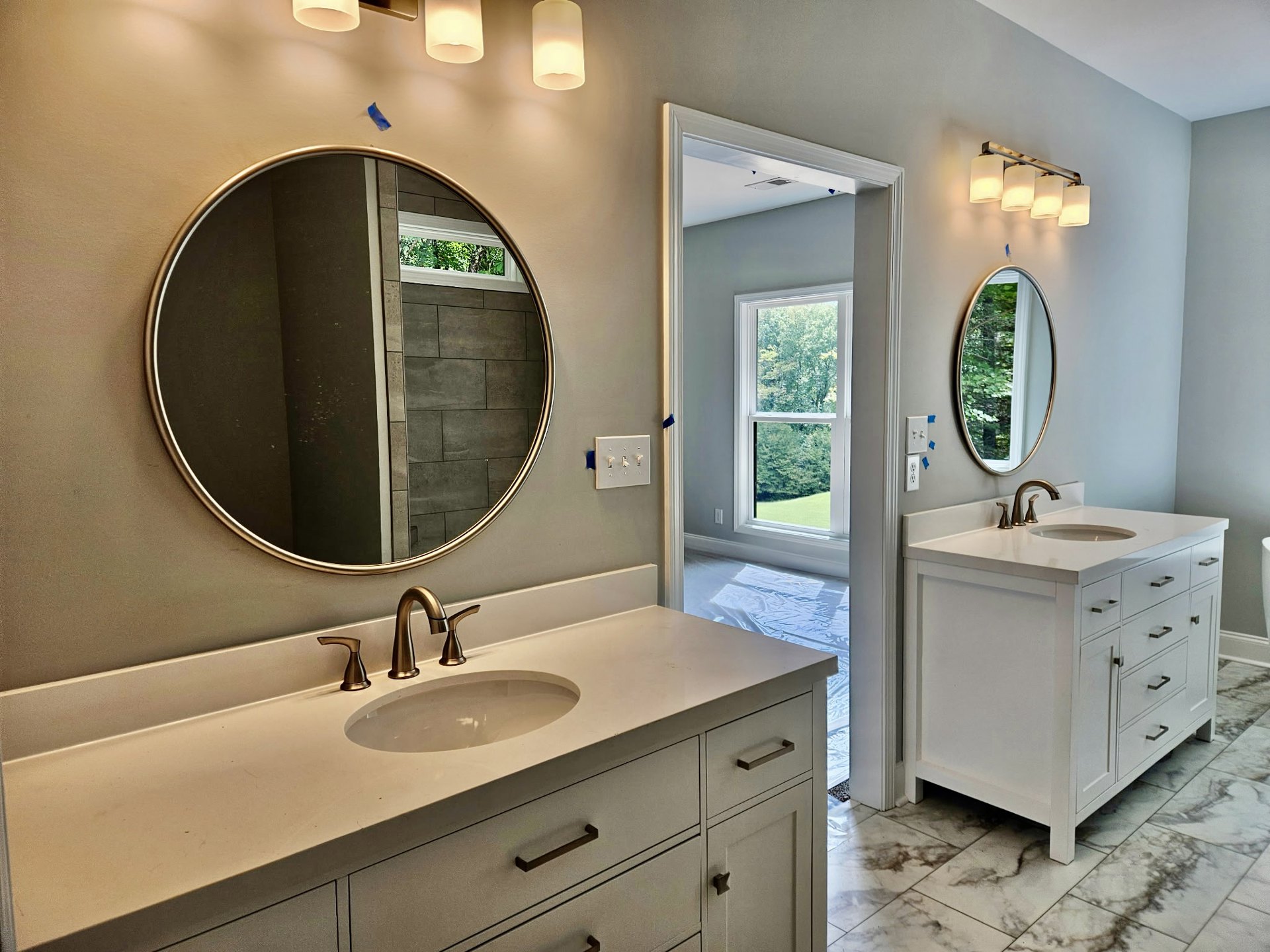 Modern bathroom with rectangular mirror above white sink, chrome faucet, tiled backsplash, light switch panel, and window revealing green trees outside