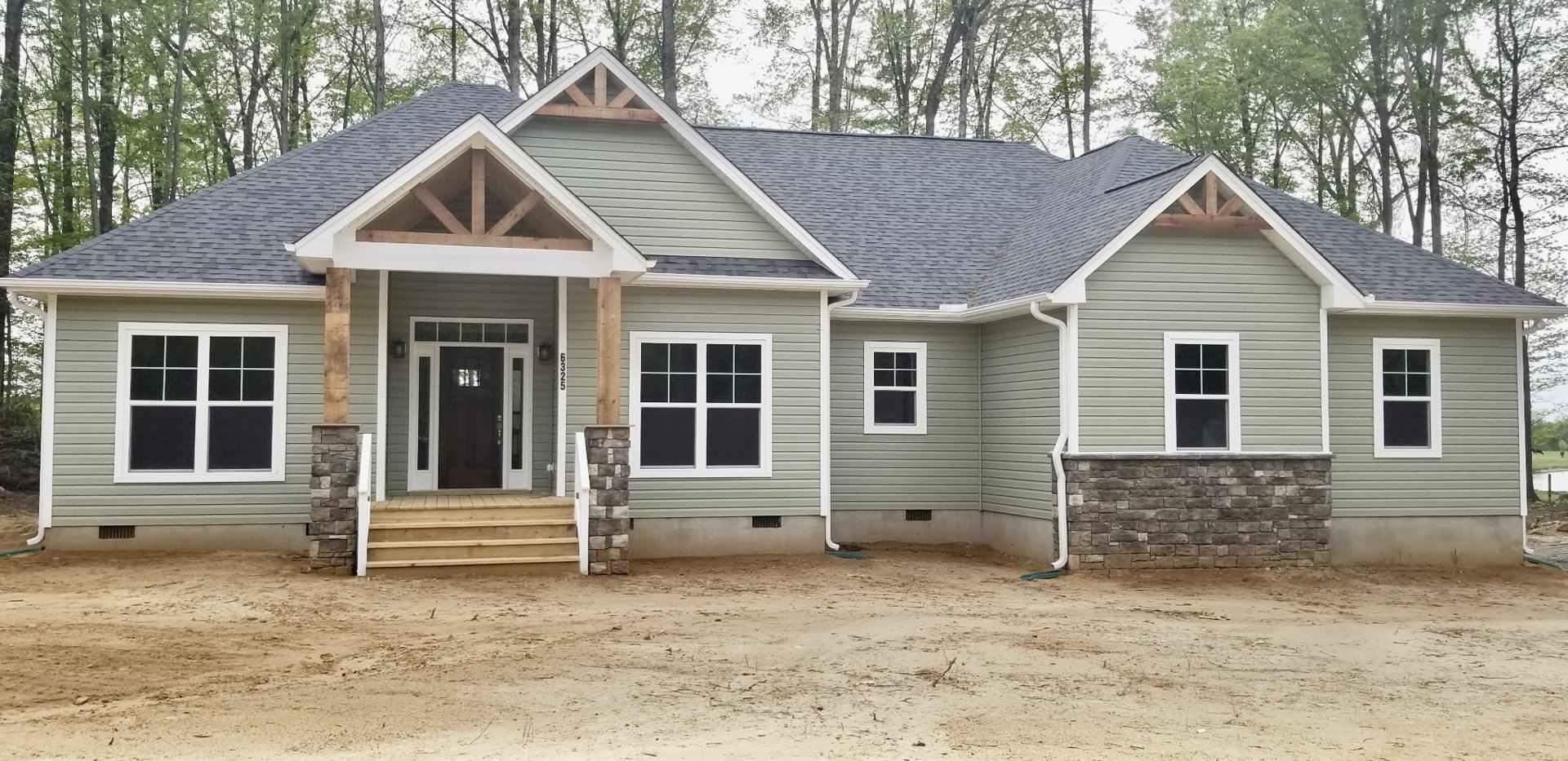 Tan siding house with white-framed windows, wooden porch and steps, dirt driveway, exposed pipe on bare ground, surrounded by trees.