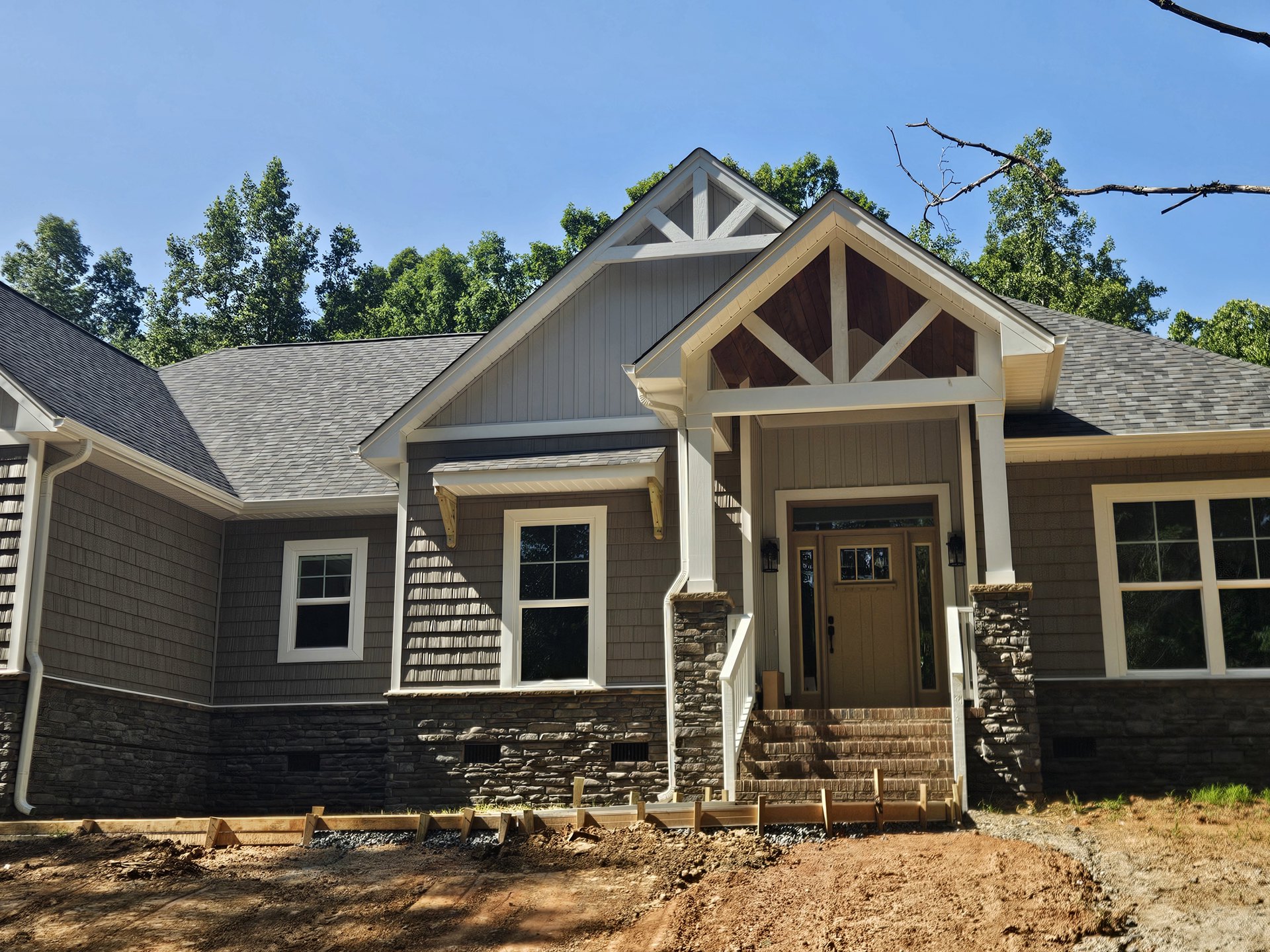 Two-story home with white-framed windows, wooden front porch and stairs, glass-paneled front door, gray siding, gabled roof, and mature trees in the background