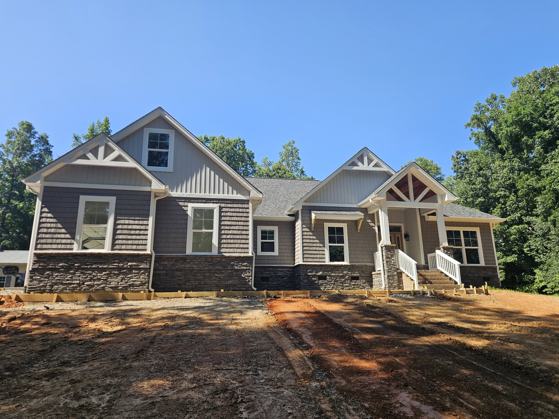 Two-story home with wood siding and stone wall, white-framed window, covered porch, dirt road in front, brick wall along driveway, mature trees and blue sky in background