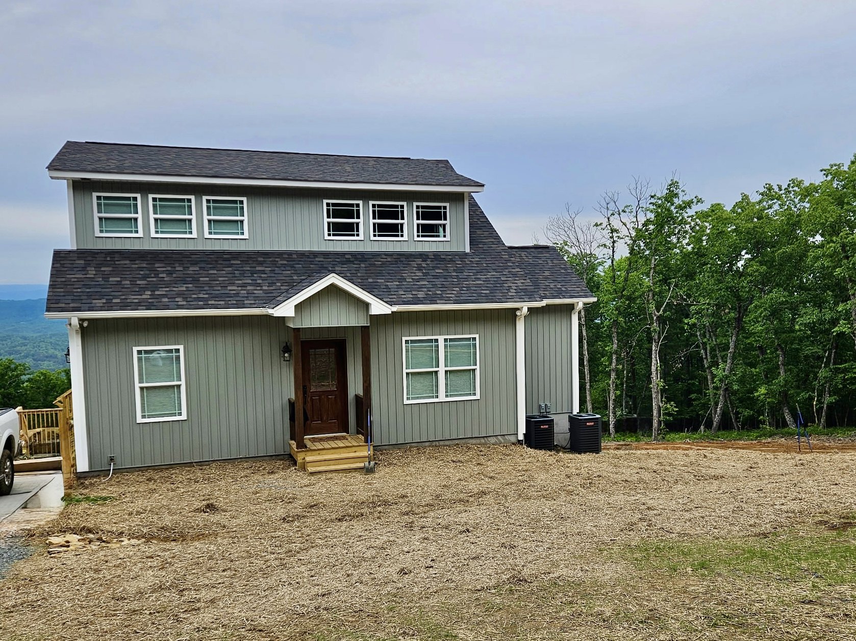 Two-story cottage-style home with white siding, front porch, large windows with blinds, wooden door, manicured grass yard, mature trees, and partly cloudy sky.