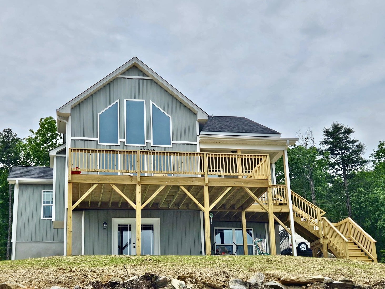 Blue-sided house with white trim, elevated wooden deck, staircase leading to yard, square-paned window, door with cracked glass, leafy trees in background.