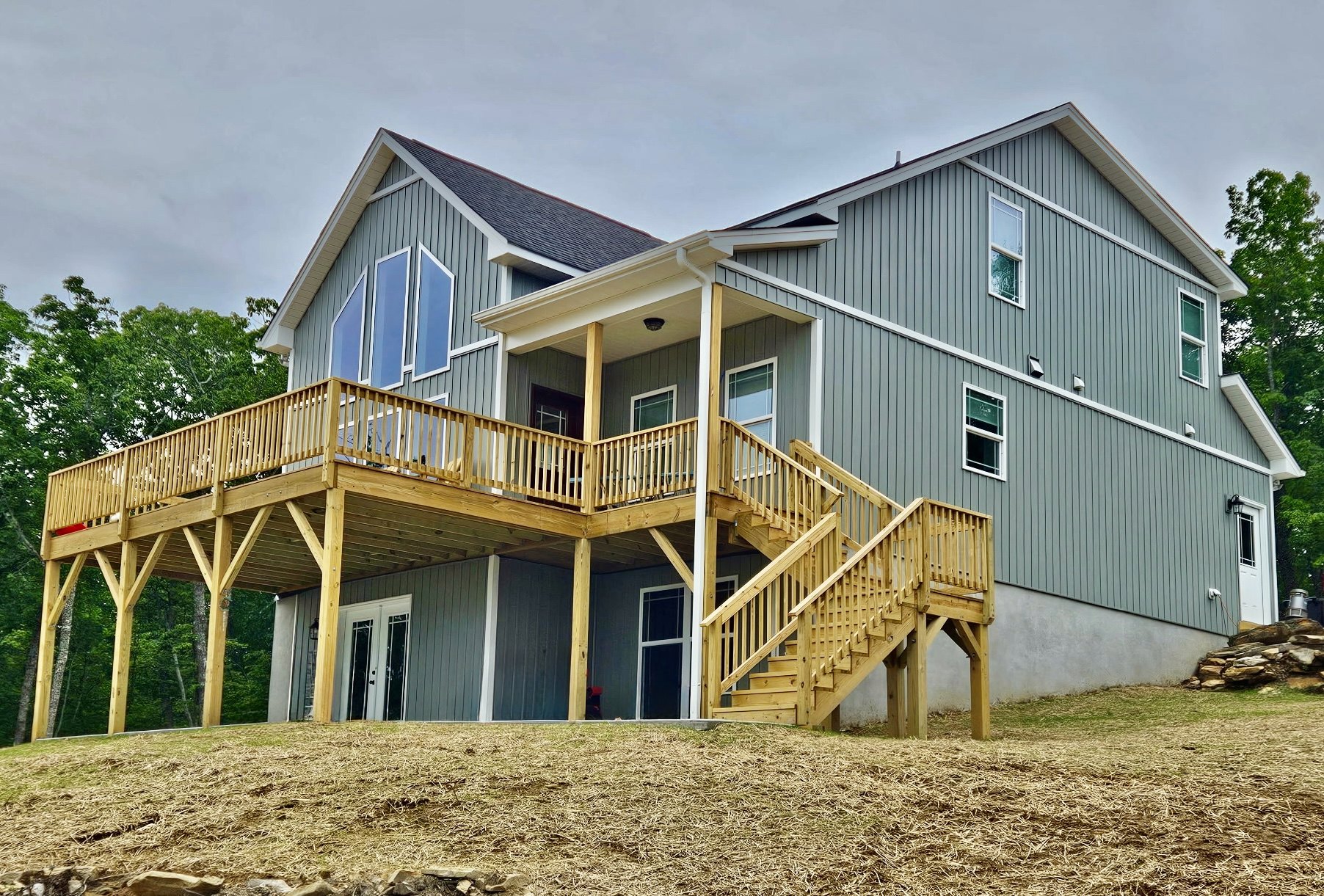 Two-story home with light siding, elevated wooden deck, white-framed windows, staircase leading to grassy yard, and mature trees in background