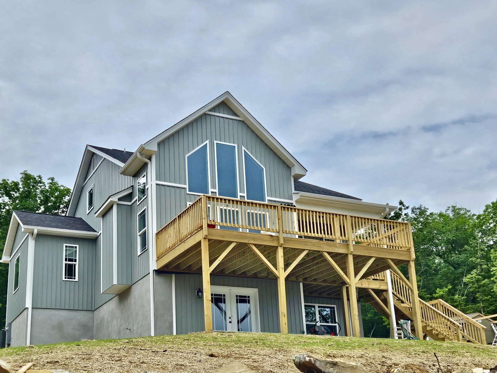 Two-story home with white siding, expansive covered porch, wooden deck, double glass doors, and white trim, surrounded by trees under a partly cloudy sky