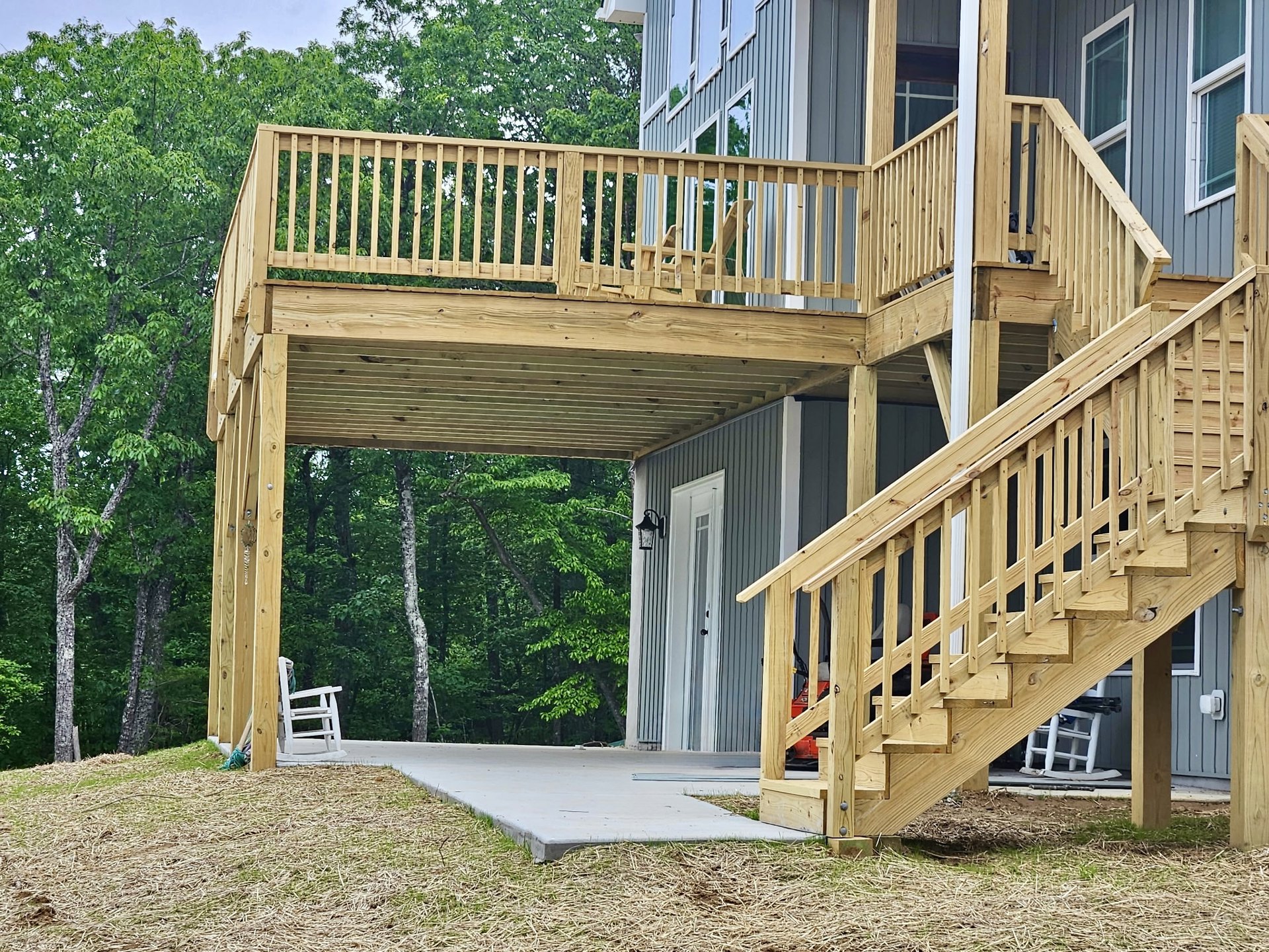 Wooden deck with stairs leading to a covered porch, white chair draped with blue scarf beside a wood table, window with planter box, surrounded by trees and greenery.