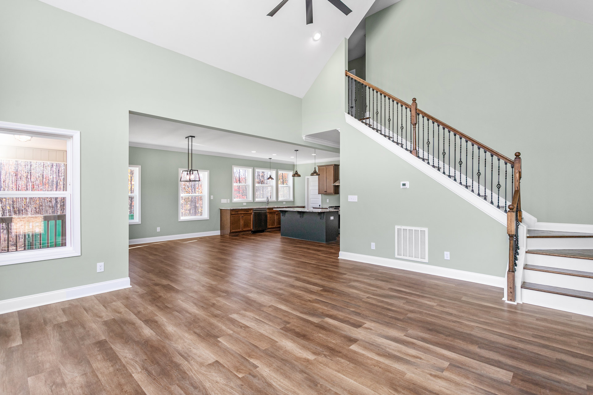 Open-concept room with hardwood flooring, ceiling fan, metal-railed staircase, large window overlooking trees, black refrigerator, and white wall vent