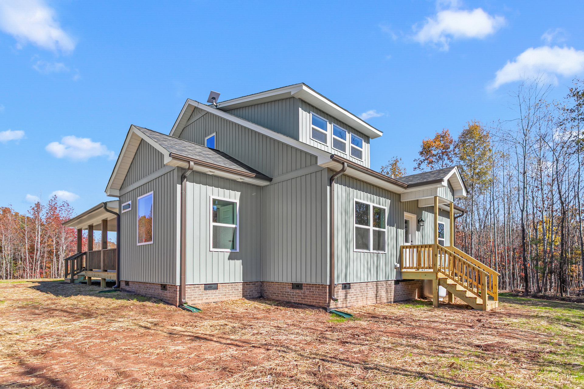 Two-story house with gray siding, white-trimmed windows, covered porch, wooden deck, and stairs leading to landscaped yard