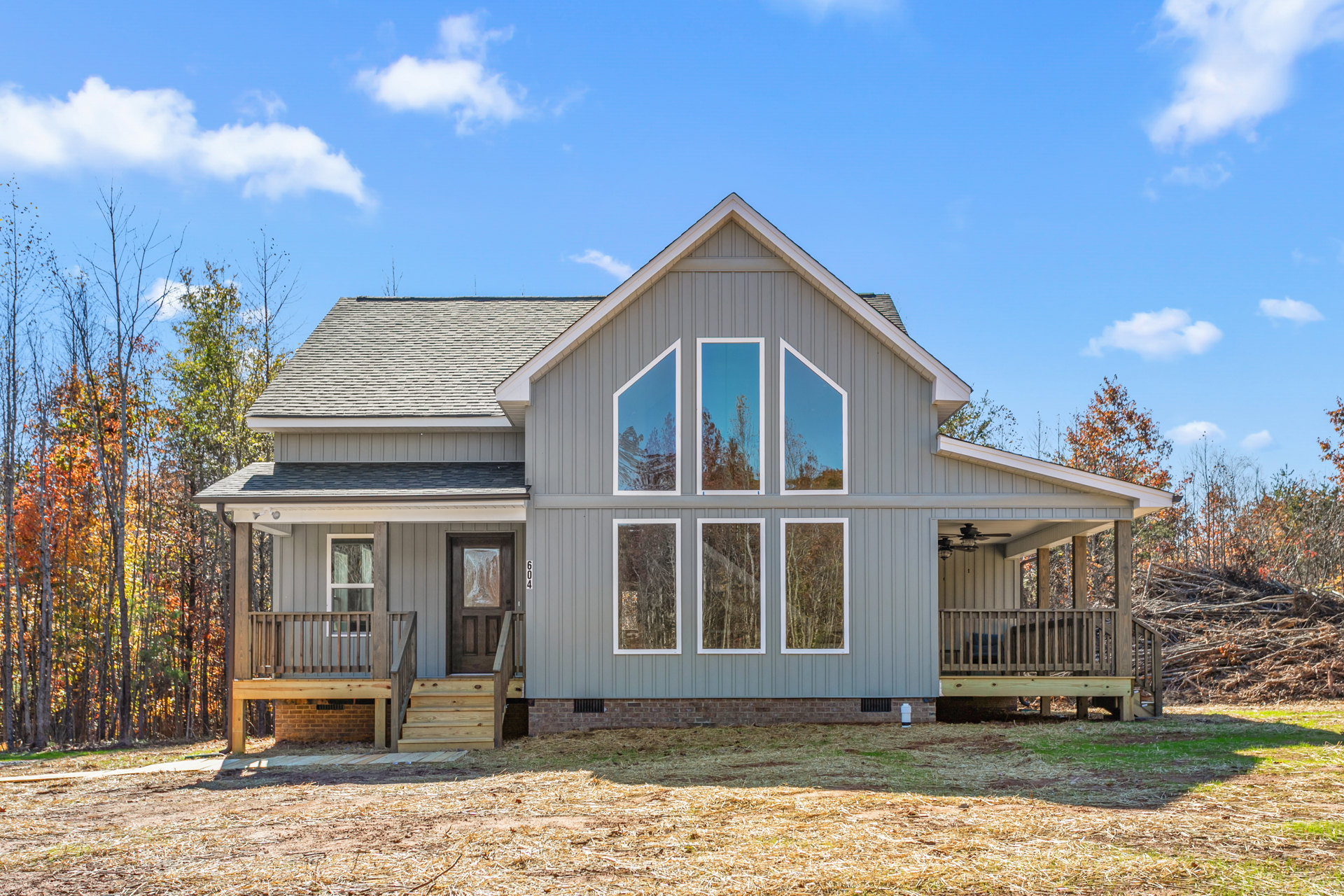 Two-story house with white siding, covered front porch, wooden deck, multiple windows reflecting blue sky and surrounding trees, landscaped yard with mature trees