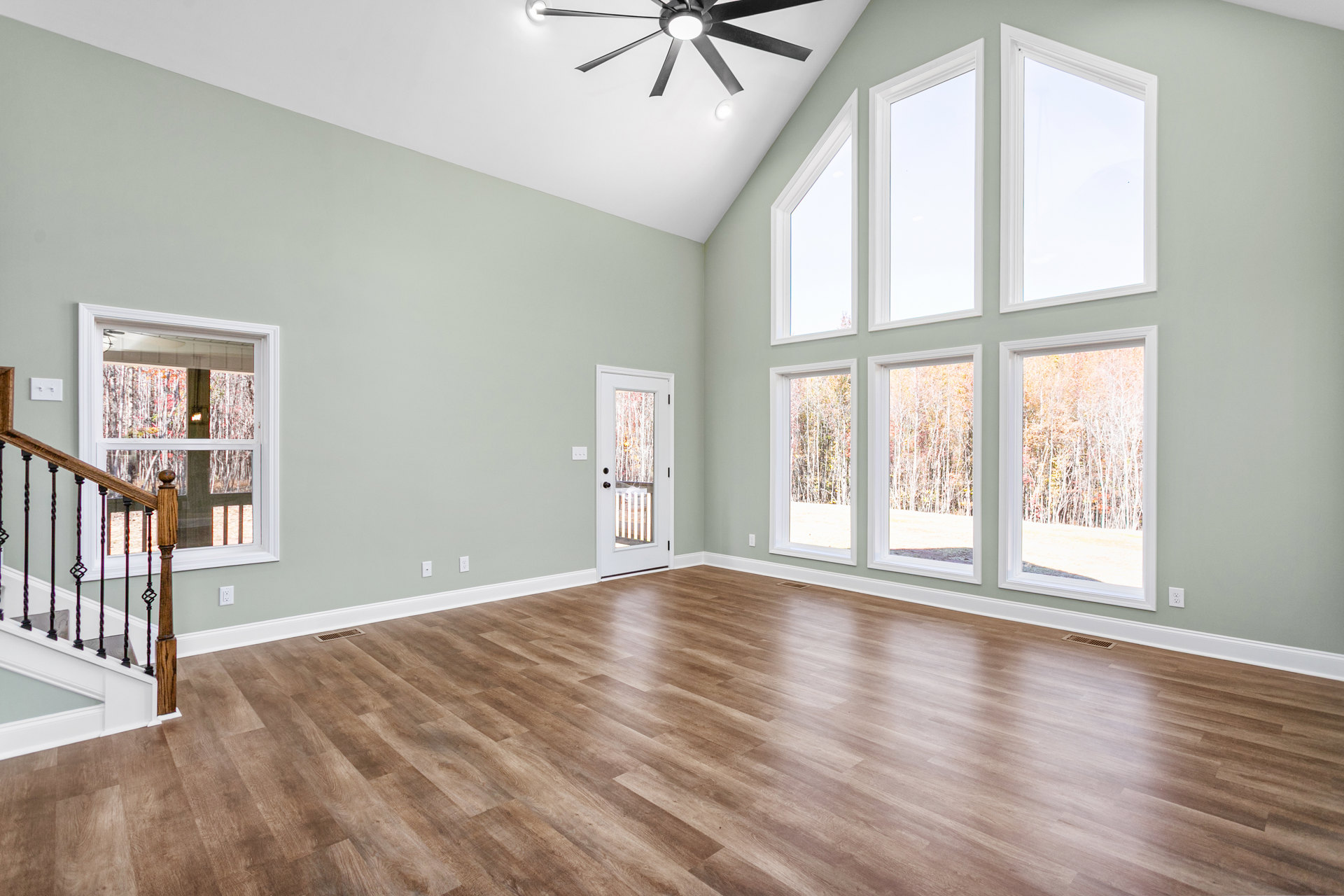 Open room with hardwood floor, white walls, ceiling fan with light, large windows showing trees outside, close-up of staircase, white door with glass panel.