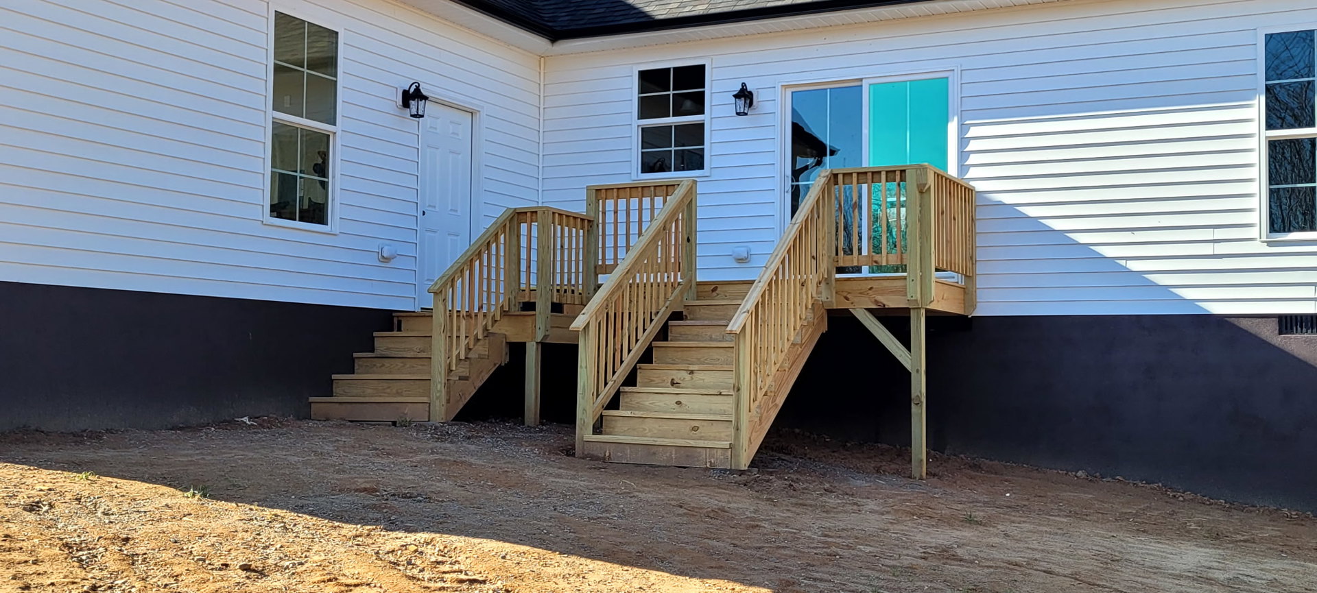 Wooden exterior stairs leading to a porch, black light fixture mounted on white siding, multi-pane window with white frame, wooden structure set on bare ground