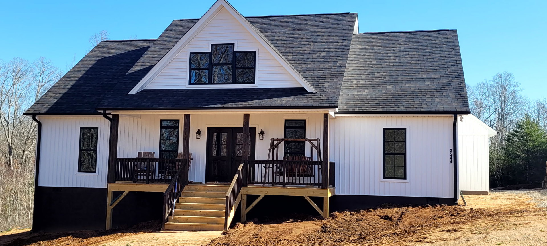 Modern home exterior featuring a wooden deck with black railing, glass-paneled door, black-framed windows reflecting trees, and outdoor wooden stairs.