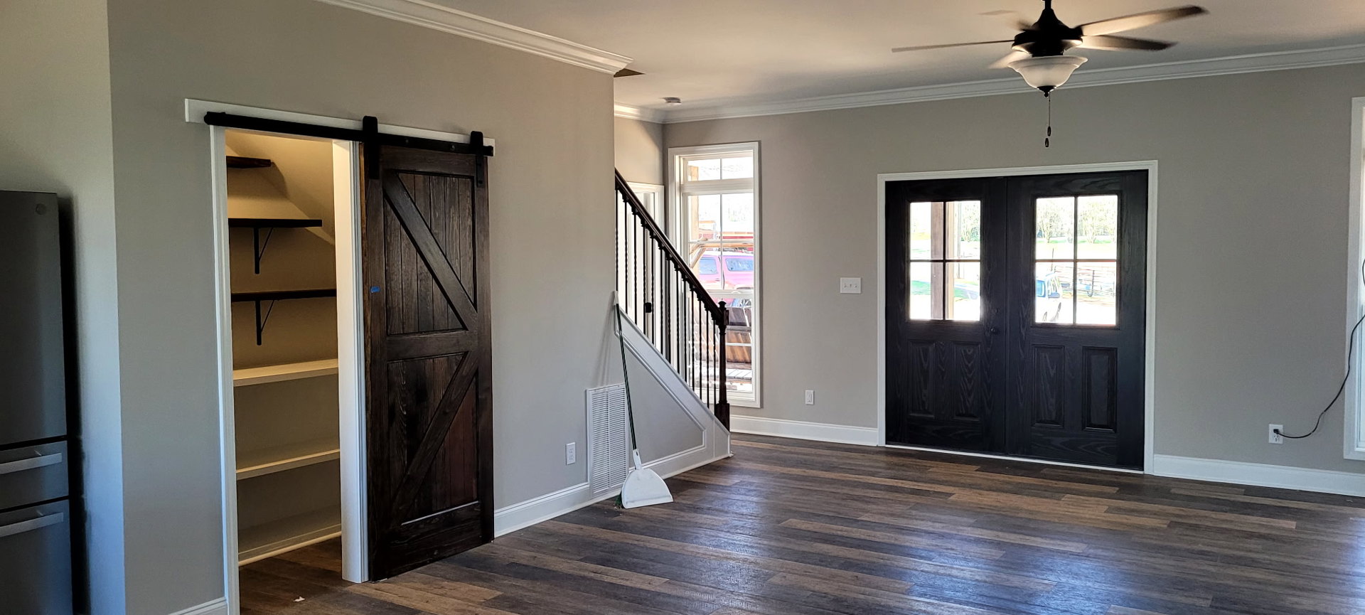 Wood flooring and white walls in a room featuring a double wooden door with glass panes, blue tape on the door, a broom leaning against the wall, and a staircase visible in the