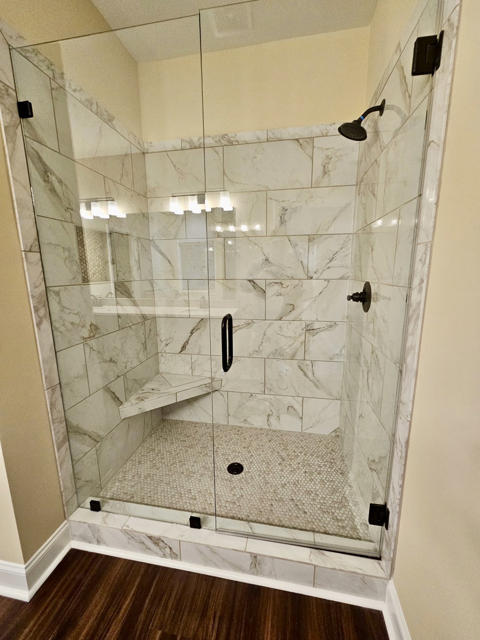 Glass shower enclosure with marble bench, white tile floor, wood flooring outside shower, black metal fixture on white plaster wall, chrome shower head.
