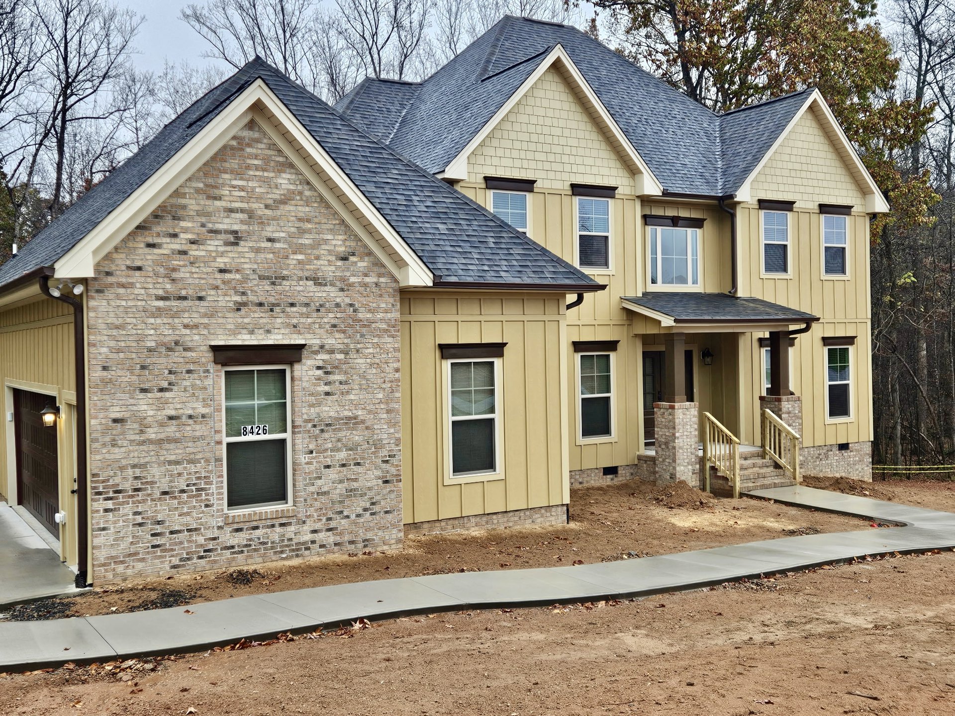 Brick and stone exterior home with white-framed windows, numbered window, concrete walkway bordered by dirt, wooden stair railings, and paved driveway.