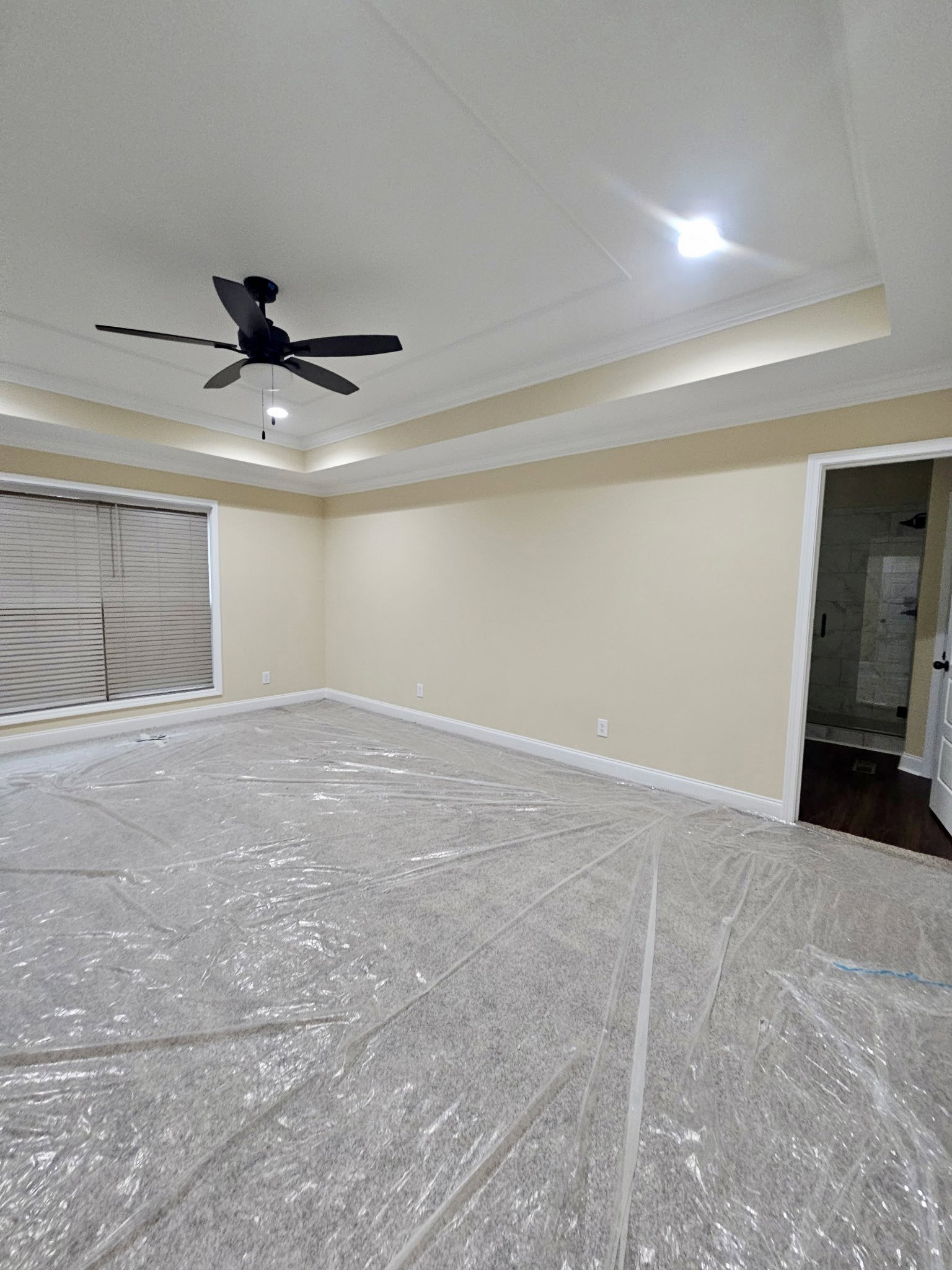 Ceiling fan with light fixture above a room featuring plastic covering on the floor, window with closed blinds, and glass bathroom door.